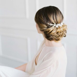May include: A woman with long brown hair styled in a low bun with a delicate floral hair comb. The comb features small white flowers and gold accents.