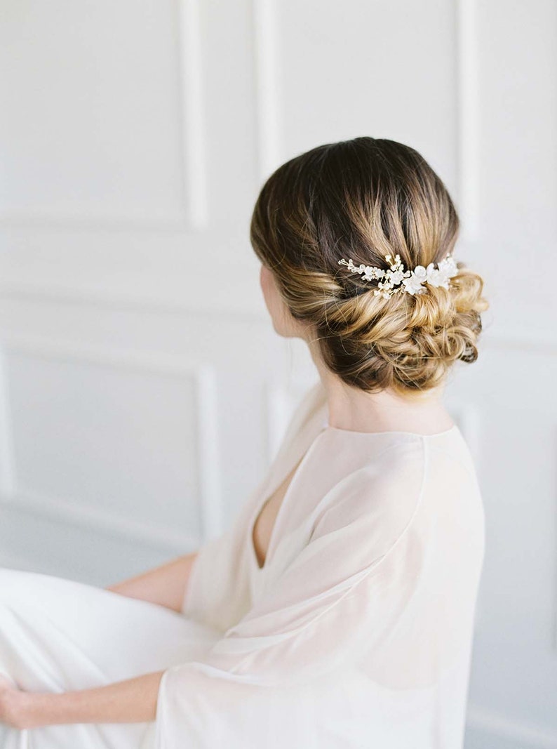 May include: A woman with long brown hair styled in a low bun with a delicate floral hair comb. The comb features small white flowers and gold accents.
