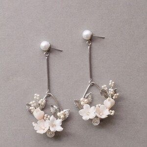 May include: A pair of silver dangle earrings with a delicate floral design. The earrings feature small white flowers and leaves, accented with tiny pearls. The earrings are suspended from a silver post with a pearl at the top.