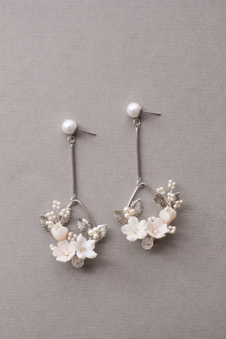 May include: A pair of silver dangle earrings with a delicate floral design. The earrings feature small white flowers and leaves, accented with tiny pearls. The earrings are suspended from a silver post with a pearl at the top.