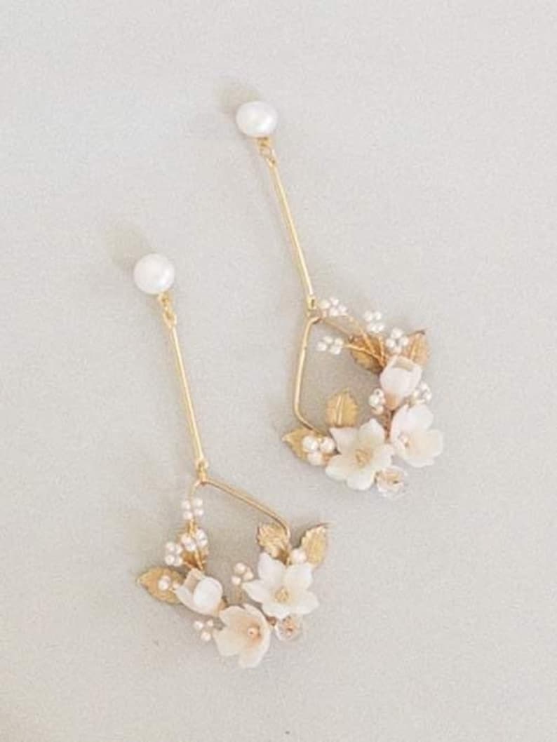 May include: A pair of gold earrings with delicate white floral accents and a pearl at the top of each earring.