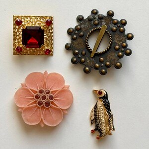 May include: A collection of four brooches: a square gold-tone piece with a large red center stone, a circular dark metal pin with a central bar, a light pink carved flower brooch with small red accents, and a small gold and black penguin pin.