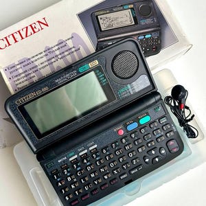 Puede incluir: Un reproductor digital Citizen ED-680 negro con teclado, altavoz y pantalla. El dispositivo está abierto, revelando la pantalla y el teclado. La caja es blanca con el logotipo de Citizen y texto en varios idiomas. Se incluye un juego de auriculares.
