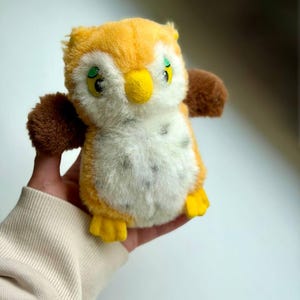 May include: A small plush owl toy featuring a golden-yellow head and back, brown wings, and a white chest with grey speckles. The owl has a bright yellow beak, yellow feet, and large green eyes with black pupils, crafted from soft, fuzzy material.
