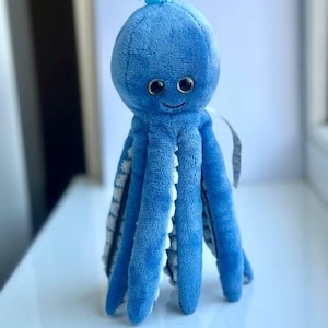 May include: A plush blue octopus toy with a smiling face and large, round eyes. The octopus has eight long tentacles with white and gray accents. A blue plastic clip is attached to the top of the toy.