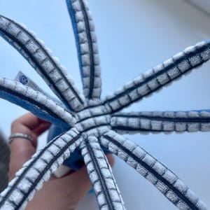 May include: A plush octopus toy with eight gray and white striped tentacles radiating from a blue central body. The tentacles have a textured, ribbed design with black accents. The toy is held against a light blue background.