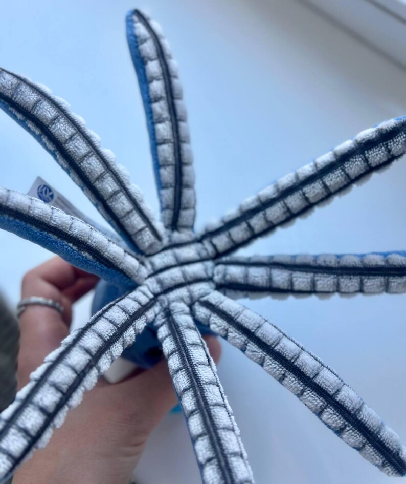 May include: A plush octopus toy with eight gray and white striped tentacles radiating from a blue central body. The tentacles have a textured, ribbed design with black accents. The toy is held against a light blue background.