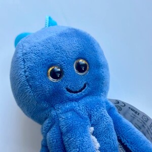 May include: A plush blue octopus toy with large, glittery eyes and a smiling mouth. The toy has eight tentacles and a small fin on top of its head. The toy is made of soft, fuzzy material.