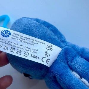 May include: A blue plush octopus toy with a white tag attached. The tag includes the text "Whitehouse Leisure International Ltd" and care instructions. The toy is designed for children aged 12 months and older.
