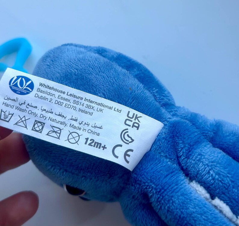 May include: A blue plush octopus toy with a white tag attached. The tag includes the text "Whitehouse Leisure International Ltd" and care instructions. The toy is designed for children aged 12 months and older.