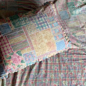 Könnte beinhalten: Ein rechteckiges Kissen und ein passendes Bettwäscheset mit Patchwork-Design, bestehend aus verschiedenen Quadraten mit Blumen-, Karomustern und Streifen in sanften Rosa-, Blau-, Grün- und Gelbtönen. Das Kissen hat einen Rüschenrand.