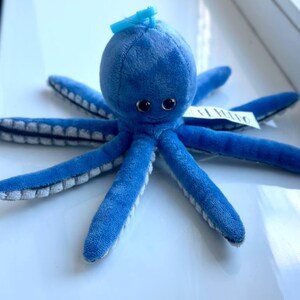 May include: A plush blue octopus toy with eight long tentacles. The octopus has a round head, large black eyes, and a small smile. The tentacles are a lighter blue on the underside. A small tag is attached to one of the tentacles.