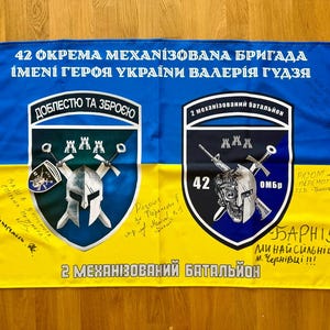 May include: A blue and yellow flag with Ukrainian text and military insignia. The flag features the text "42 OKPEMA MEXAHI3OBANA БРИГАДА" and "2 МЕХАНІЗОВАНИЙ БАТАЛЬЙОН". The flag has two crests with a helmet and crossed weapons.