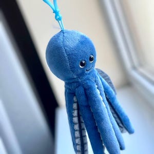 May include: A plush blue octopus keychain with a smiling face and eight tentacles. The keychain has a light blue clip attached to the top of the octopus's head. The tentacles have a gray and white pattern on the underside.