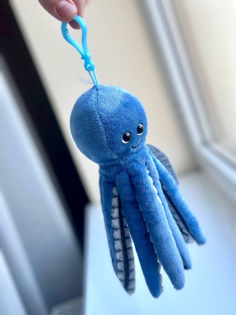 May include: A plush blue octopus keychain with a smiling face and eight tentacles. The keychain has a light blue clip attached to the top of the octopus's head. The tentacles have a gray and white pattern on the underside.