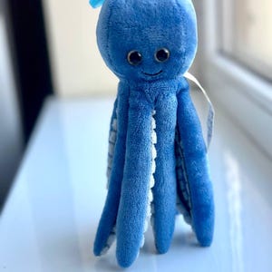 May include: A plush blue octopus toy with a smiling face and large, dark eyes. The tentacles have white scalloped edges. A loop of blue fabric is attached to the top of the head. The toy is sitting on a white surface.