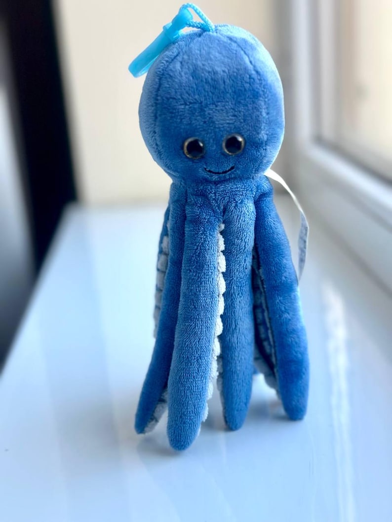 May include: A plush blue octopus toy with a smiling face and large, dark eyes. The tentacles have white scalloped edges. A loop of blue fabric is attached to the top of the head. The toy is sitting on a white surface.