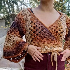 May include: A crocheted wrap top in shades of brown, tan, and purple. The top features a V-neckline, long bell sleeves, and a tie closure at the waist. The open weave design adds a delicate touch to this handmade garment.