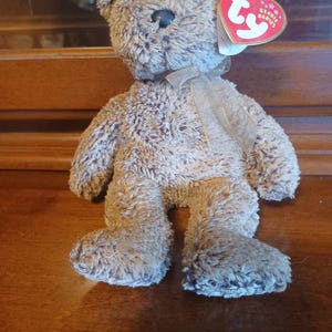 May include: A brown plush teddy bear with a Ty heart-shaped tag. The bear has a light brown ribbon around its neck and a black nose. The bear is standing on a wooden surface.