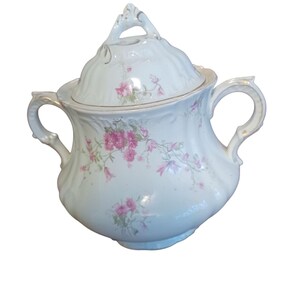 May include: A white porcelain sugar bowl with a lid, adorned with pink floral patterns and gold trim. The bowl has two handles and a decorative finial on the lid. A vintage piece, perfect for serving sugar.
