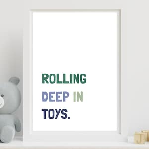 May include: A framed print with the words "ROLLING DEEP IN TOYS" in green, blue, and navy. A gray teddy bear and wooden blocks are on the shelf below the print. A small potted plant is to the right of the print.