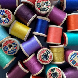 Può includere: Una collezione di rocchetti di filo colorati in varie tonalità di viola, blu, arancione, giallo e verde. I rocchetti hanno i lati in legno e sono etichettati con testo tra cui "National" e "100 YARDS".