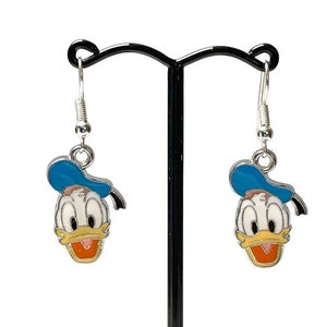Puede incluir: Pendientes colgantes con la cara de Donald Duck. Cada pendiente tiene un gancho plateado y un dije de esmalte colorido que representa la cara de Donald Duck, con un sombrero azul y un pico naranja.