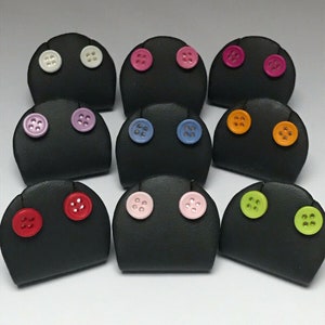 以下が含まれることがあります： 9つの黒いイヤリングディスプレイスタンドのコレクション。それぞれにカラフルなボタンが2つずつ付いています。ボタンは白、ピンク、青、オレンジ、緑など様々な色があり、遊び心のあるデザインです。