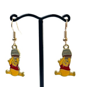 May include: Gold-coloured dangle earrings featuring a yellow cartoon bear in a red shirt, holding a honey pot. The honey pot is brown with the word "HUNNY" printed on it. The earrings are displayed on a black earring stand.