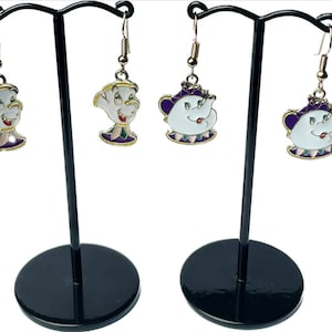 May include: Two pairs of earrings with cartoon character designs. One pair shows a white teacup with a smiling face, and the other pair depicts a white teapot with a smiling face. Both pairs have gold-coloured hooks and are displayed on black stands.