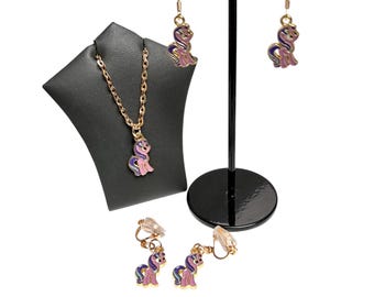 May include: A jewelry set featuring a gold-tone chain necklace with a pony pendant, a pair of gold-tone hook earrings with matching pony charms, and a pair of gold-tone clip-on earrings with matching pony charms, all with purple and pink enamel.