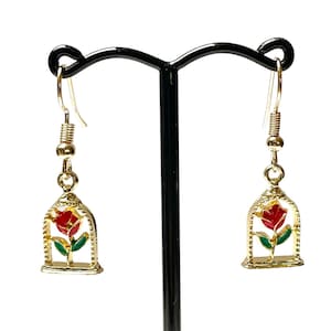 May include: Gold-colored dangle earrings featuring a red rose inside a gold cage design. The rose has green leaves and is set against a white background. The earrings are displayed on a black earring stand.