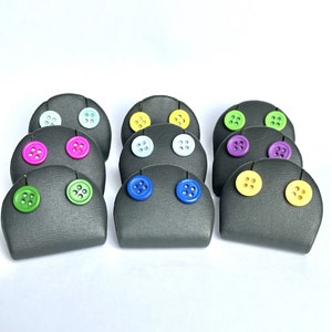 May include: A collection of button earrings displayed on gray earring holders. The earrings feature colorful buttons in various shades, including pink, green, blue, and yellow. The background is plain white.