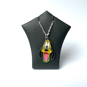 May include: A silver chain necklace with a cartoon dog pendant. The pendant is yellow, black, and pink, with a green collar. The necklace is displayed on a black jewelry stand against a white background.