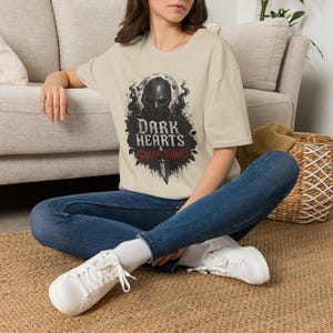 May include: A beige short-sleeve t-shirt featuring a graphic print of a dark masked figure surrounded by roses and smoke. The text reads Dark Hearts & Sharp Things, with a dagger illustration positioned below the text in black and red ink.