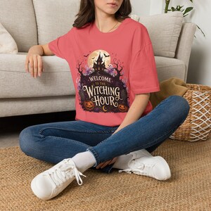 May include: A coral-colored short-sleeve t-shirt featuring a graphic print of a haunted house, a full moon, bats, and pumpkins. The text reads Welcome to the Witching Hour in a decorative font. The shirt is paired with blue denim jeans and white sneakers.