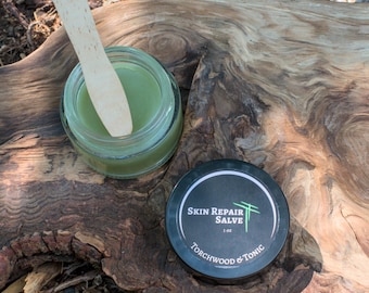 May include: A jar of green salve with a wooden applicator rests on a piece of weathered wood. A black container with the text "Skin Repair Salve" and "Torchwood & Tonic" is also present. The items are set on a bed of leaves and twigs.