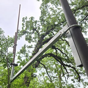 May include: A metal antenna support system attached to a dark pole with a U-bolt. A long, thin antenna extends upwards, and a horizontal metal bar is secured to the pole. The background features lush green trees and a cloudy sky.