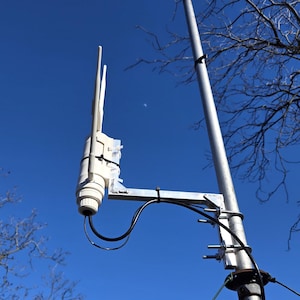 May include: A white outdoor wireless access point with two antennas is mounted on a metal pole. The device is secured with black zip ties and connected to black cables. The background is a clear blue sky.