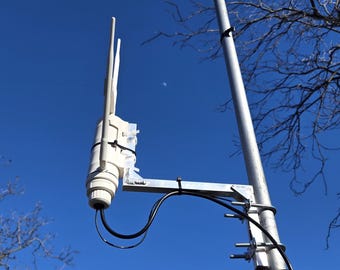 Puede incluir: Un punto de acceso inalámbrico para exteriores blanco con dos antenas está montado en un poste de metal. El dispositivo está asegurado con bridas negras y conectado a cables negros. El fondo es un cielo azul claro.