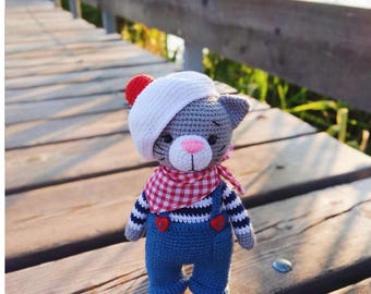 May include: A handmade crochet cat doll wearing a white beret with a red pom-pom, a red and white checkered scarf, and blue overalls with red heart-shaped buttons. The cat has gray fur, a pink nose, and is standing on a wooden surface.