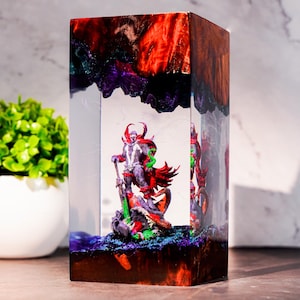 May include: A rectangular resin block containing a miniature Spawn figure. The top and bottom sections feature swirling red, orange, and purple patterns. The central clear section displays the character with a red cape and green accents.