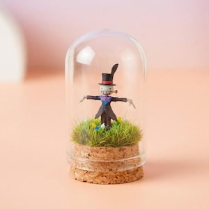 May include: A miniature figurine of a scarecrow character inside a glass dome. The figure wears a black top hat, a purple scarf, and a dark coat. It stands on a bed of green grass and small flowers, set on a cork base.