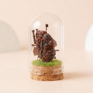 May include: A miniature brown steampunk-style castle sculpture inside a glass dome. The sculpture sits on a bed of green faux grass, with a cork base. The dome is clear, and the background is a soft beige.