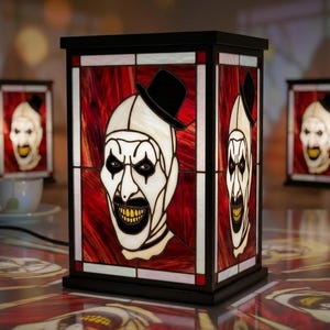 Può includere: Una lampada rettangolare in vetro colorato raffigurante un clown dal volto bianco con un cilindro nero su uno sfondo rosso vorticoso. La lampada ha una cornice nera e mostra il volto del clown su più lati, illuminata dall'interno.