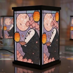 Pode incluir: Um candeeiro de mesa retangular com uma moldura preta e painéis translúcidos. Os painéis apresentam uma ilustração gráfica de uma figura escura encapuzada a segurar uma taça, rodeada por flores de cerejeira cor-de-rosa, lanternas cor de laranja e uma lua grande.