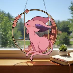 May include: A pink stained glass window hanging featuring a cartoon Espeon character. The artwork is set within a gold-colored circular frame, adorned with pink floral accents and a chain for hanging. The background is a view of a sunny day.