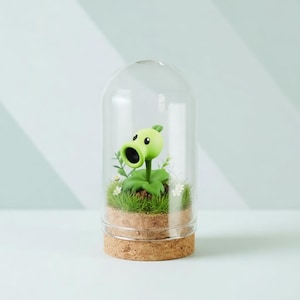 May include: A decorative terrarium featuring a green cartoon pea shooter figure. The figure is inside a clear glass dome, set on a cork base. The base contains artificial grass, small white flowers, and greenery.
