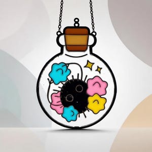 Peut inclure: Ornement en forme de bouteille en verre transparent, avec un contour noir, rempli de formes florales colorées et d'une sphère noire. La bouteille a un bouchon marron et est suspendue par une chaîne. Les fleurs sont bleues, roses et jaunes.