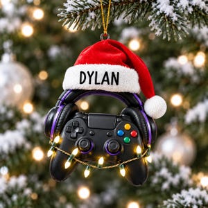 May include: A black video game controller ornament wearing a red and white Santa hat with the name DYLAN printed on the white brim. The controller is adorned with purple accents, black headphones resting on top, and a string of small glowing Christmas lights.